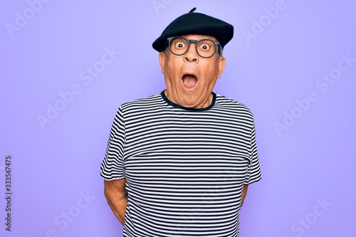 Senior handsome hoary man wearing french beret and glasses standing over purple background afraid and shocked with surprise expression, fear and excited face.