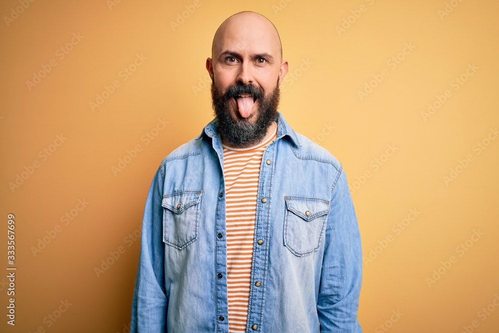 Handsome bald man with beard wearing casual denim jacket and striped t-shirt sticking tongue out happy with funny expression. Emotion concept. Handsome bald man with beard wearing casual denim jacket and striped t-shirt sticking tongue out happy with funny expression. Emotion concept.