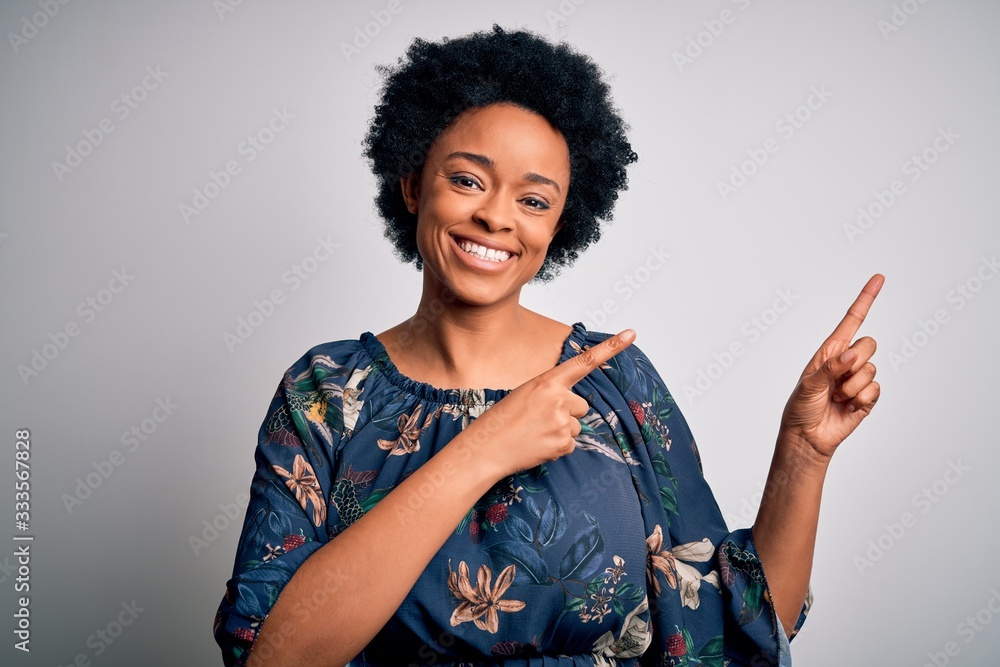 Young beautiful African American afro woman with curly hair wearing casual floral dress smiling and looking at the camera pointing with two hands and fingers to the side. Young beautiful African American afro woman with curly hair wearing casual floral dress smiling and looking at the camera pointing with two hands and fingers to the side.