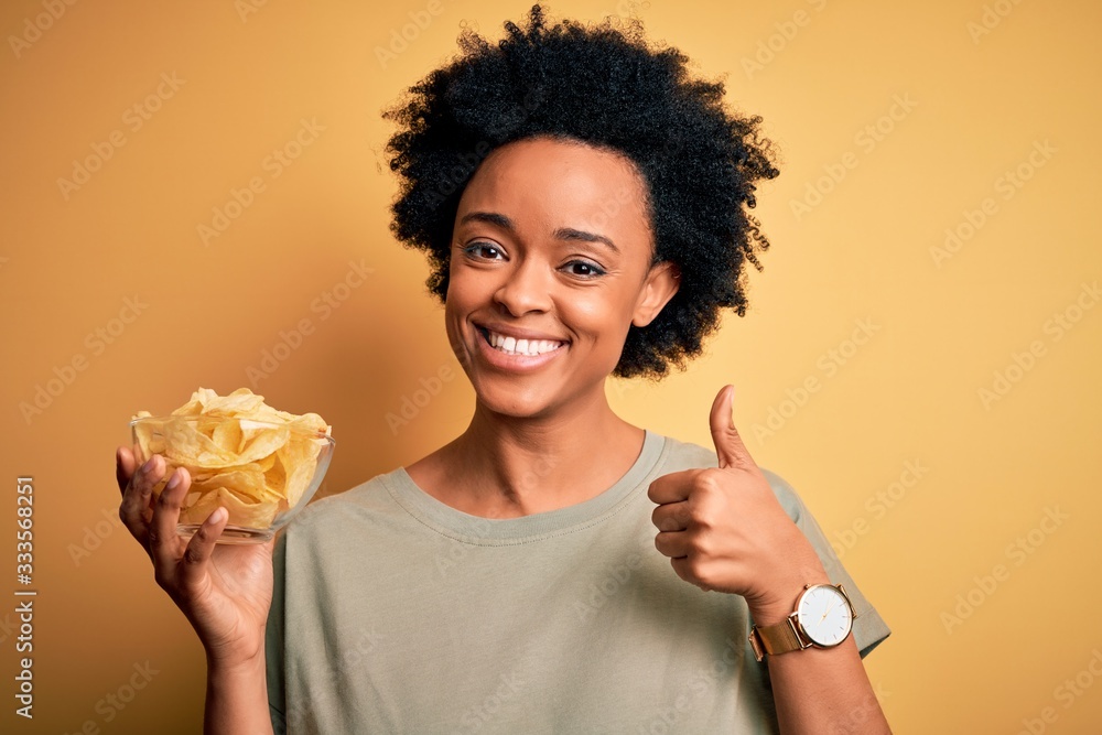 Young African American afro woman with curly hair holding bowl with chips potatoes happy with big smile doing ok sign, thumb up with fingers, excellent sign Young African American afro woman with curly hair holding bowl with chips potatoes happy with big smile doing ok sign, thumb up with fingers, excellent sign