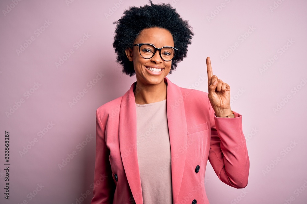 Young beautiful African American afro businesswoman with curly hair wearing pink jacket showing and pointing up with finger number one while smiling confident and happy. Young beautiful African American afro businesswoman with curly hair wearing pink jacket showing and pointing up with finger number one while smiling confident and happy.