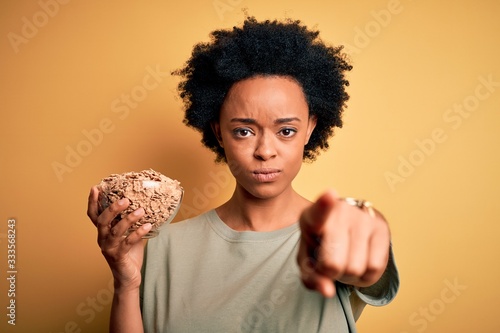 Young African American afro woman with curly hair holding bowl with cornflakes cereals pointing with finger to the camera and to you  hand sign  positive and confident gesture from the front