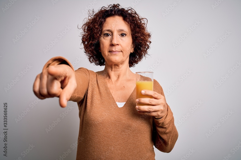 Middle age curly hair woman drinking healthy glass of orange juice over white background pointing with finger to the camera and to you, hand sign, positive and confident gesture from the front Middle age curly hair woman drinking healthy glass of orange juice over white background pointing with finger to the camera and to you, hand sign, positive and confident gesture from the front