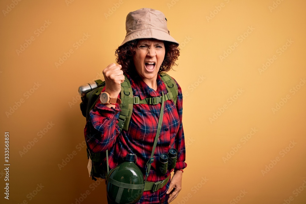 Middle age curly hair hiker woman hiking wearing backpack and water canteen using binoculars angry and mad raising fist frustrated and furious while shouting with anger. Rage and aggressive concept.