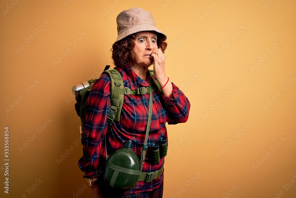 Middle age curly hair hiker woman hiking wearing backpack and water canteen using binoculars looking stressed and nervous with hands on mouth biting nails. Anxiety problem.