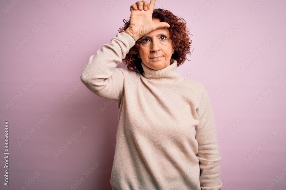 Middle age beautiful curly hair woman wearing casual turtleneck sweater over pink background making fun of people with fingers on forehead doing loser gesture mocking and insulting. Middle age beautiful curly hair woman wearing casual turtleneck sweater over pink background making fun of people with fingers on forehead doing loser gesture mocking and insulting.