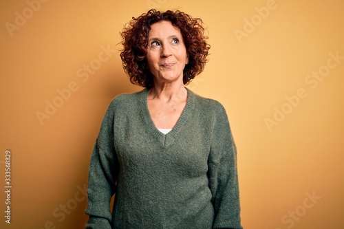Middle age beautiful curly hair woman wearing casual sweater over isolated yellow background smiling looking to the side and staring away thinking. © Krakenimages.com