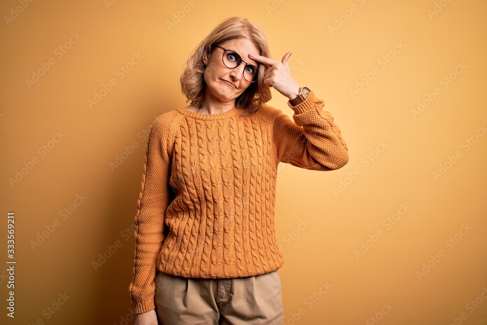 Middle age beautiful blonde woman wearing casual sweater and glasses over yellow background pointing unhappy to pimple on forehead, ugly infection of blackhead. Acne and skin problem Middle age beautiful blonde woman wearing casual sweater and glasses over yellow background pointing unhappy to pimple on forehead, ugly infection of blackhead. Acne and skin problem