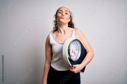 Young beautiful woman with curly hair holding weighing machine over white background looking at the camera blowing a kiss on air being lovely and sexy. Love expression.
