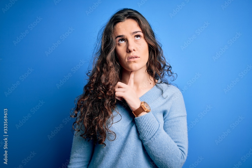 Fototapeta premium Young beautiful woman with curly hair wearing blue casual sweater over isolated background Thinking concentrated about doubt with finger on chin and looking up wondering