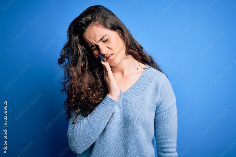 Young beautiful woman with curly hair wearing blue casual sweater over isolated background touching mouth with hand with painful expression because of toothache or dental illness on teeth. Dentist Young beautiful woman with curly hair wearing blue casual sweater over isolated background touching mouth with hand with painful expression because of toothache or dental illness on teeth. Dentist