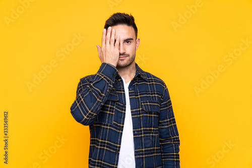 Young handsome man over isolated yellow background covering a eye by hand