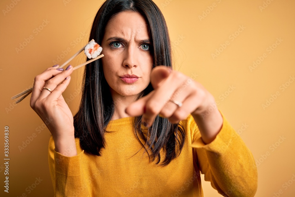 Obraz premium Young brunette woman with blue eyes eating salmon maki sushi using chopsticks pointing with finger to the camera and to you, hand sign, positive and confident gesture from the front