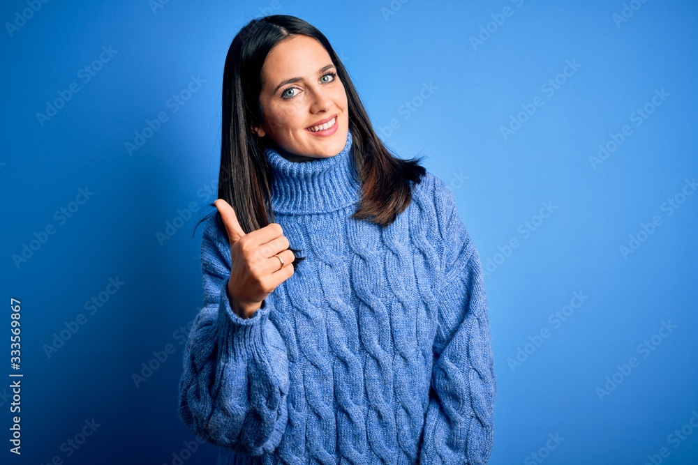 Young brunette woman with blue eyes wearing casual turtleneck sweater doing happy thumbs up gesture with hand. Approving expression looking at the camera showing success. Young brunette woman with blue eyes wearing casual turtleneck sweater doing happy thumbs up gesture with hand. Approving expression looking at the camera showing success.