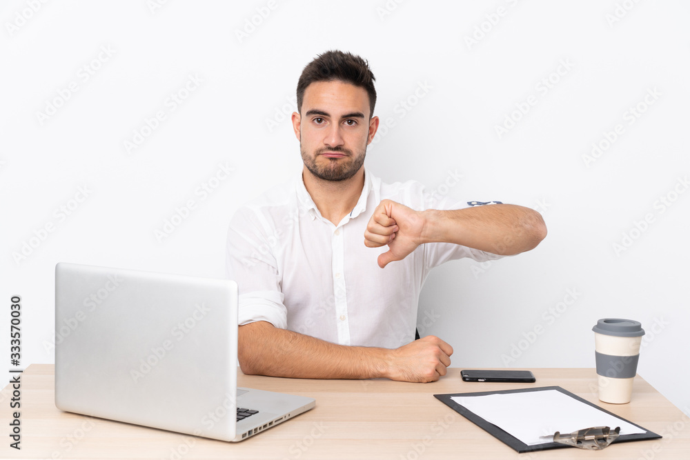 Young business man with a mobile phone in a workplace showing thumb down sign Young business man with a mobile phone in a workplace showing thumb down sign