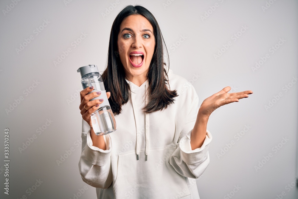 Young brunette sporty woman with blue eyes holding water bottle over white background very happy and excited, winner expression celebrating victory screaming with big smile and raised hands Young brunette sporty woman with blue eyes holding water bottle over white background very happy and excited, winner expression celebrating victory screaming with big smile and raised hands