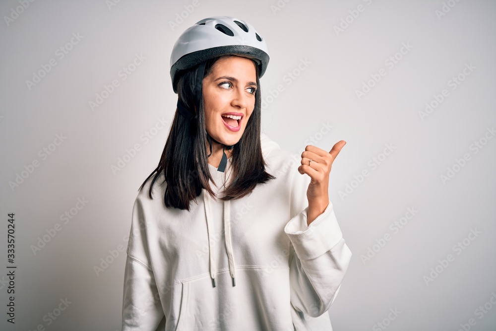 Young cyclist woman with blue eyes wearing bike helmet over isolated white background smiling with happy face looking and pointing to the side with thumb up.