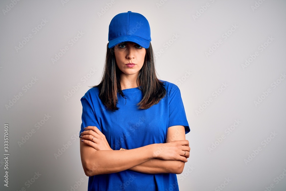 Young delivery woman with blue eyes wearing cap standing over blue background skeptic and nervous, disapproving expression on face with crossed arms. Negative person.