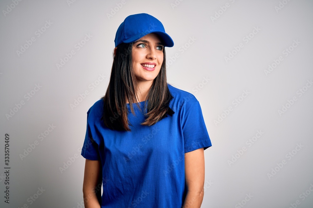 Young delivery woman with blue eyes wearing cap standing over blue background looking away to side with smile on face, natural expression. Laughing confident. Young delivery woman with blue eyes wearing cap standing over blue background looking away to side with smile on face, natural expression. Laughing confident.