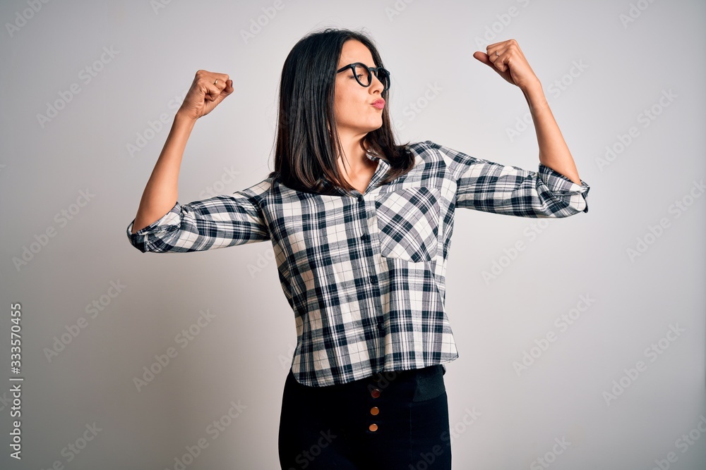 Young brunette woman with blue eyes wearing casual shirt and glasses over white background showing arms muscles smiling proud. Fitness concept. Young brunette woman with blue eyes wearing casual shirt and glasses over white background showing arms muscles smiling proud. Fitness concept.