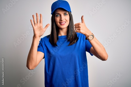 Young delivery woman with blue eyes wearing cap standing over blue background showing and pointing up with fingers number six while smiling confident and happy.