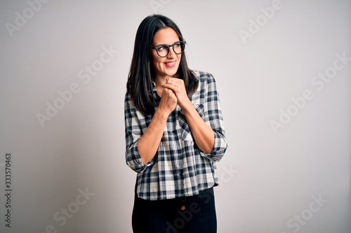 Young brunette woman with blue eyes wearing casual shirt and glasses over white background laughing nervous and excited with hands on chin looking to the side