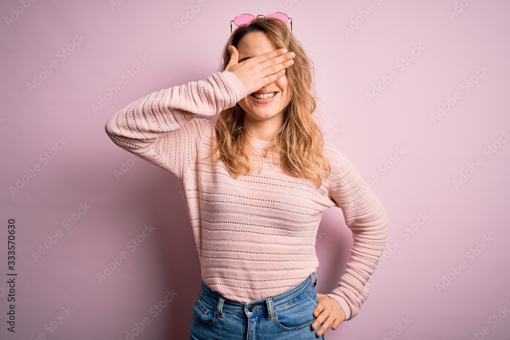 Young beautiful blonde woman wearing casual sweater and sunglasses over pink background smiling and laughing with hand on face covering eyes for surprise. Blind concept. Young beautiful blonde woman wearing casual sweater and sunglasses over pink background smiling and laughing with hand on face covering eyes for surprise. Blind concept.