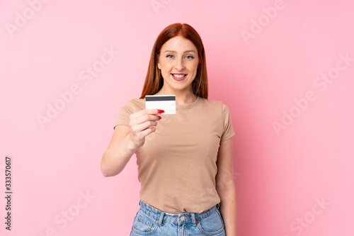 Young redhead woman over isolated pink background holding a credit card