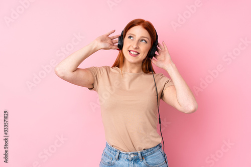 Young redhead woman over isolated pink background using the mobile with headphones