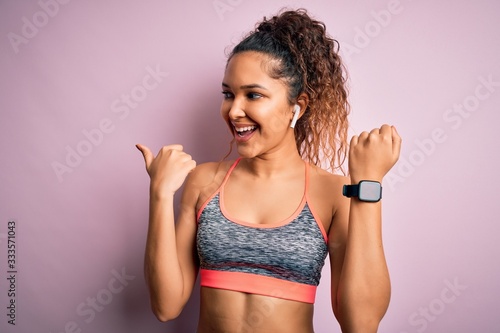 Beautiful sportswoman with curly hair doing sport wearing sportswear using smartwatch pointing and showing with thumb up to the side with happy face smiling © Krakenimages.com
