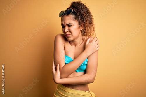 Young beautiful american woman on vacation wearing bikini over isolated yellow background shaking and freezing for winter cold with sad and shock expression on face