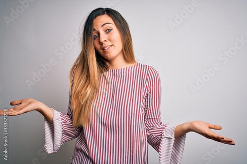 Young beautiful blonde woman with blue eyes wearing stiped t-shirt over white background clueless and confused expression with arms and hands raised. Doubt concept.