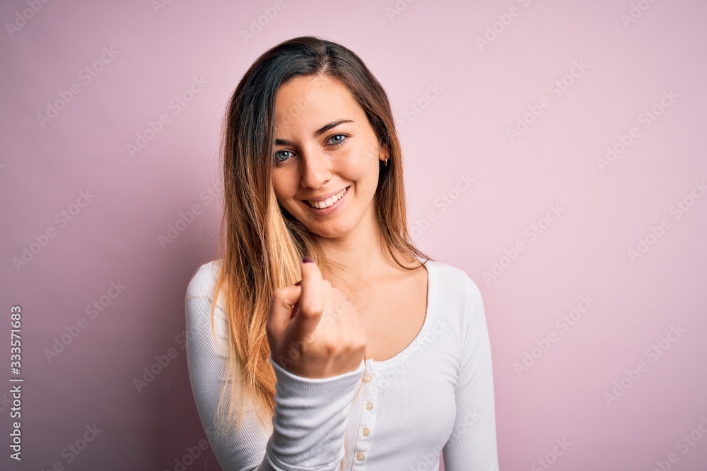 Young beautiful blonde woman with blue eyes wearing white t-shirt over pink background Beckoning come here gesture with hand inviting welcoming happy and smiling Young beautiful blonde woman with blue eyes wearing white t-shirt over pink background Beckoning come here gesture with hand inviting welcoming happy and smiling