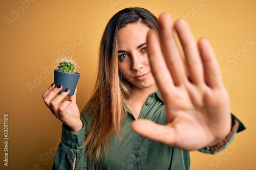 Young beautiful blonde woman with blue eyes holding small cactus pot over yellow bckground with open hand doing stop sign with serious and confident expression, defense gesture