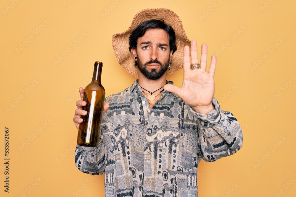 Young handsome hispanic bohemian hipster man with bear drinking from a empty wine bottle with open hand doing stop sign with serious and confident expression, defense gesture Young handsome hispanic bohemian hipster man with bear drinking from a empty wine bottle with open hand doing stop sign with serious and confident expression, defense gesture
