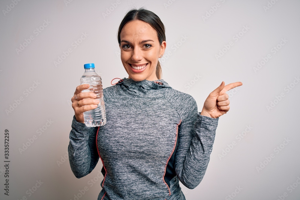 Young fitness woman wearing sport workout clothes drinking water from plastic bottle very happy pointing with hand and finger to the side Young fitness woman wearing sport workout clothes drinking water from plastic bottle very happy pointing with hand and finger to the side