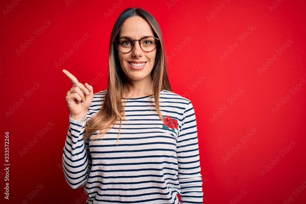 Young beautiful blonde woman with blue eyes wearing glasses standing over red background with a big smile on face, pointing with hand finger to the side looking at the camera. Young beautiful blonde woman with blue eyes wearing glasses standing over red background with a big smile on face, pointing with hand finger to the side looking at the camera.