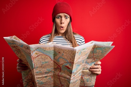 Young blonde tourist woman using holding destination city map over red background scared in shock with a surprise face, afraid and excited with fear expression