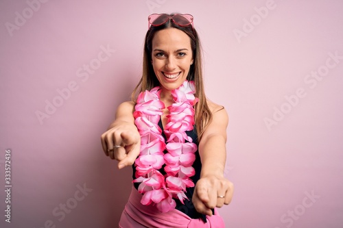 Young beautiful blonde woman wearing swimsuit and floral Hawaiian lei over pink background pointing to you and the camera with fingers  smiling positive and cheerful