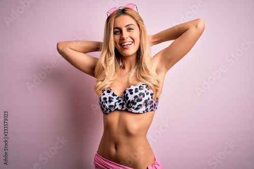 Young beautiful blonde woman on vacation wearing bikini over isolated pink background relaxing and stretching, arms and hands behind head and neck smiling happy © Krakenimages.com