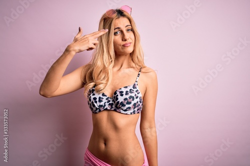 Young beautiful blonde woman on vacation wearing bikini over isolated pink background Shooting and killing oneself pointing hand and fingers to head like gun, suicide gesture. © Krakenimages.com