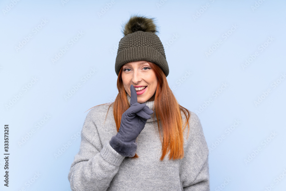 Young redhead Russian woman with winter hat over isolated blue background doing silence gesture Young redhead Russian woman with winter hat over isolated blue background doing silence gesture