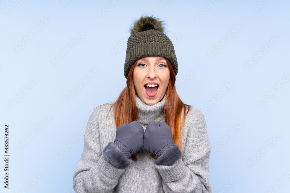 Young redhead Russian woman with winter hat over isolated blue background celebrating a victory Young redhead Russian woman with winter hat over isolated blue background celebrating a victory