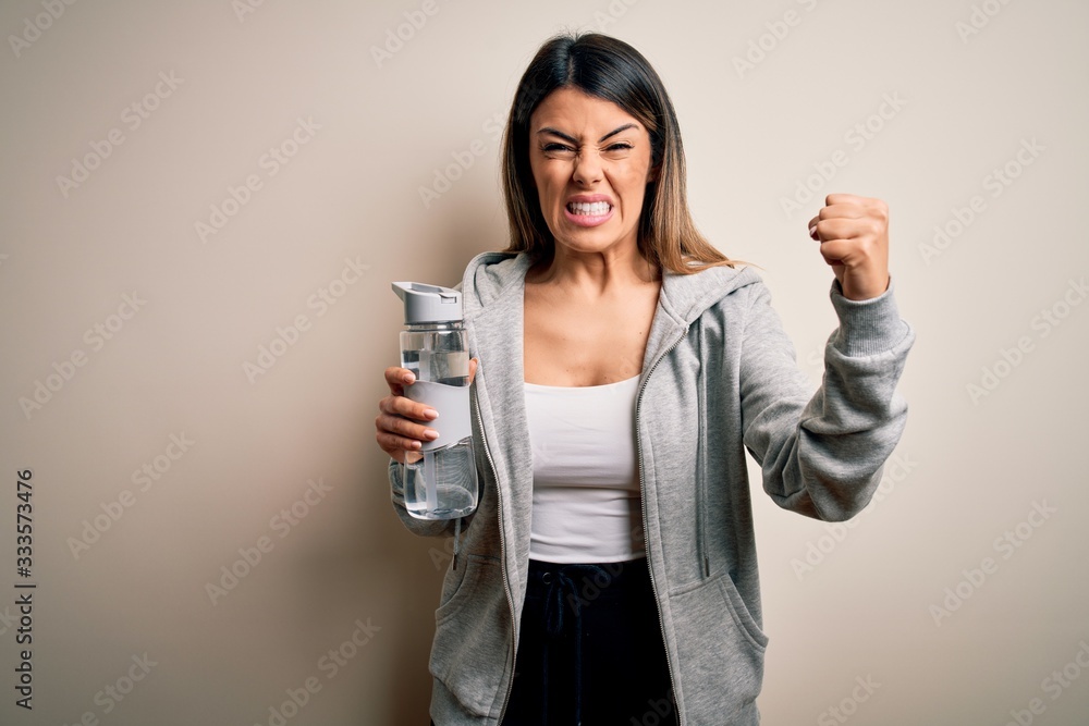 Young beautiful brunette sporty woman drinking bottle of water over isolated white background annoyed and frustrated shouting with anger, crazy and yelling with raised hand, anger concept Young beautiful brunette sporty woman drinking bottle of water over isolated white background annoyed and frustrated shouting with anger, crazy and yelling with raised hand, anger concept