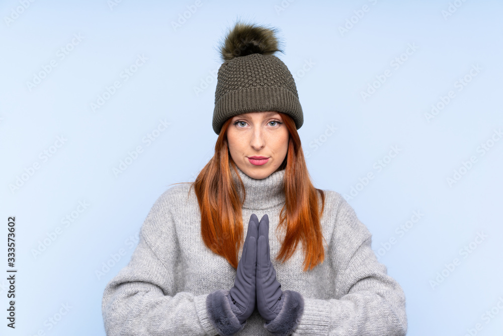 Young redhead Russian woman with winter hat over isolated blue background pleading