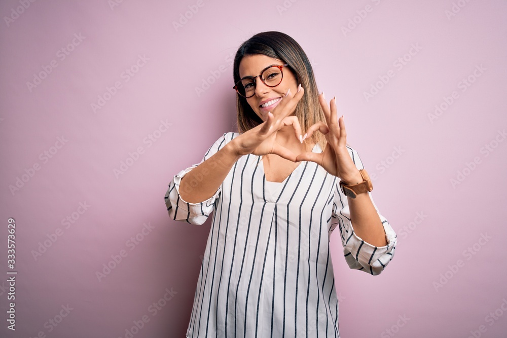 Young beautiful woman wearing casual striped t-shirt and glasses over pink background smiling in love doing heart symbol shape with hands. Romantic concept.