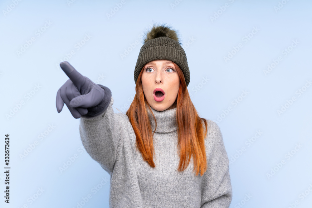 Young redhead Russian woman with winter hat over isolated blue background pointing away Young redhead Russian woman with winter hat over isolated blue background pointing away