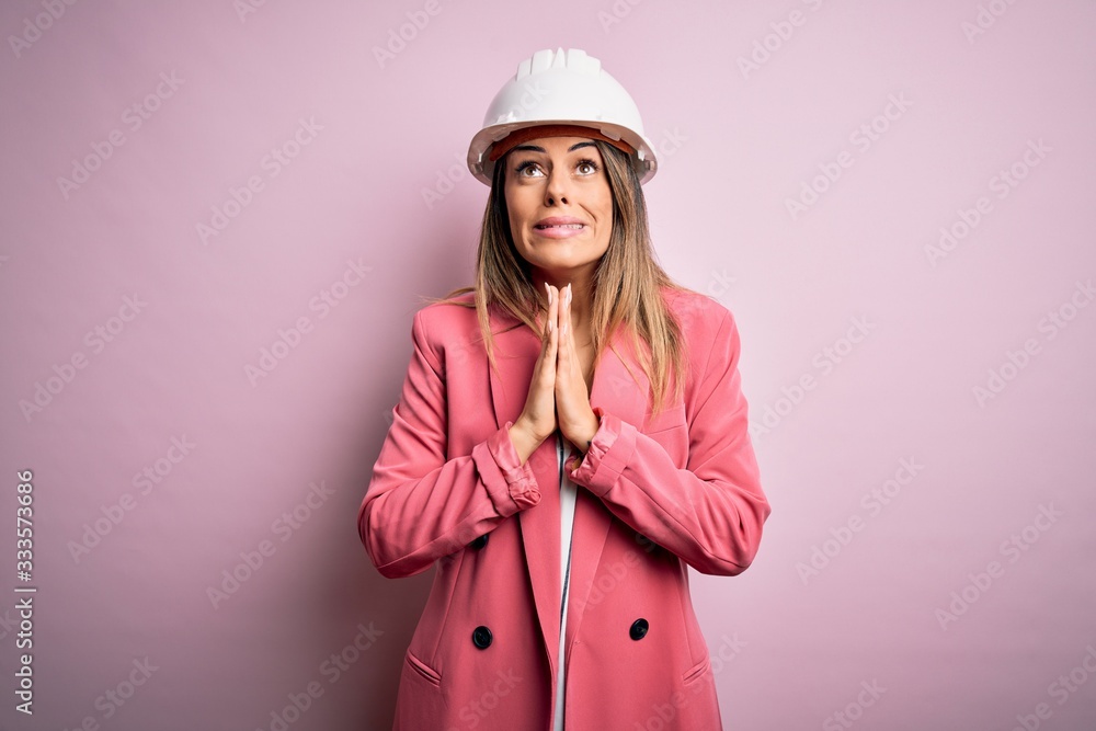 Young beautiful brunette architect woman wearing safety helmet over pink background begging and praying with hands together with hope expression on face very emotional and worried. Begging. Young beautiful brunette architect woman wearing safety helmet over pink background begging and praying with hands together with hope expression on face very emotional and worried. Begging.