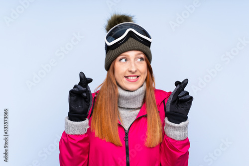 Skier redhead woman with snowboarding glasses over isolated blue wall with fingers crossing