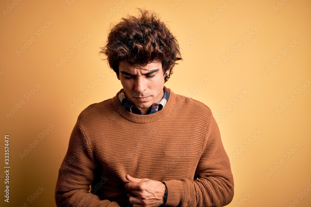 Young handsome man wearing casual shirt and sweater over isolated yellow background with hand on stomach because indigestion, painful illness feeling unwell. Ache concept. Young handsome man wearing casual shirt and sweater over isolated yellow background with hand on stomach because indigestion, painful illness feeling unwell. Ache concept.